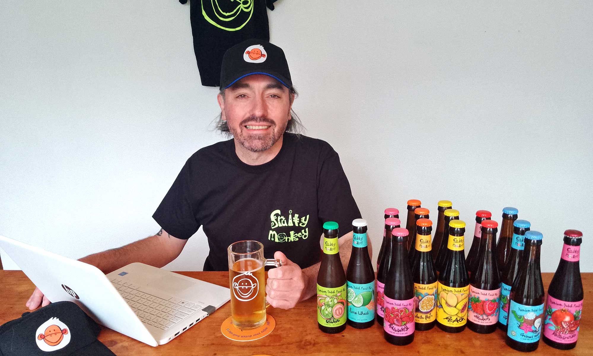 Smiling person wearing a monkey t-shirt and cap sitting at desk with laptop and beer bottles
