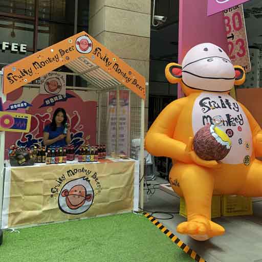 A beer stall at a festival labelled Fruity Monkey Beer, besides which sits a large yellow inflatable monkey holding a coconut and also labelled Fruity Monkey
